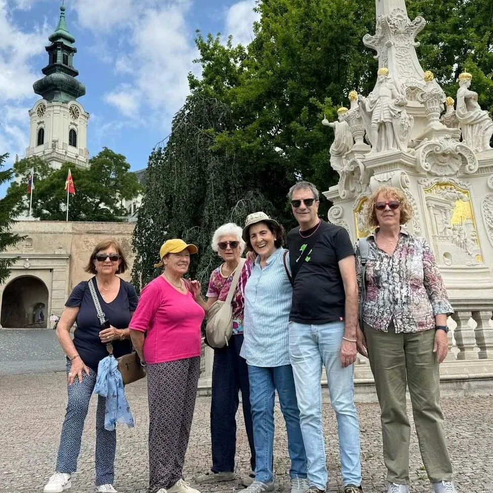 A small tour group standing in front of a baroque plague column beside a church.