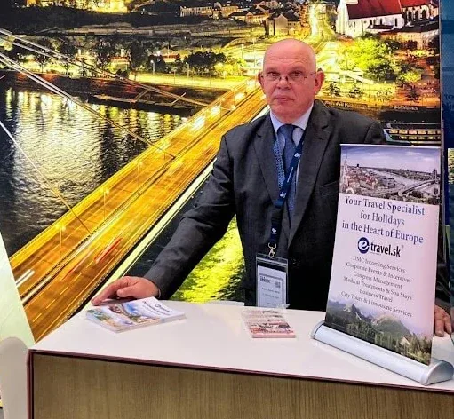 Un uomo allo stand di e-travel.sk a una fiera del turismo professionale, davanti a una grande veduta notturna di Bratislava e del Danubio.