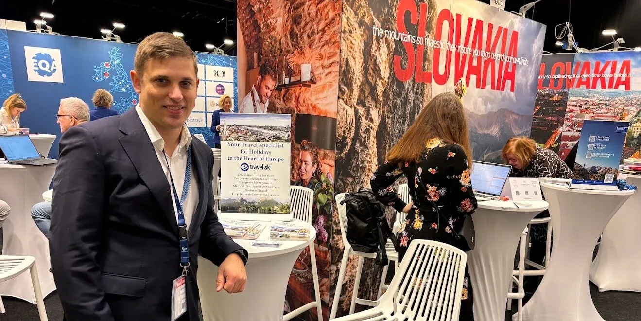 Un representante de e-travel.sk en el stand de la empresa en una feria de turismo, junto a un panel con sus servicios de DMC y un gran fondo turístico de Eslovaquia.
