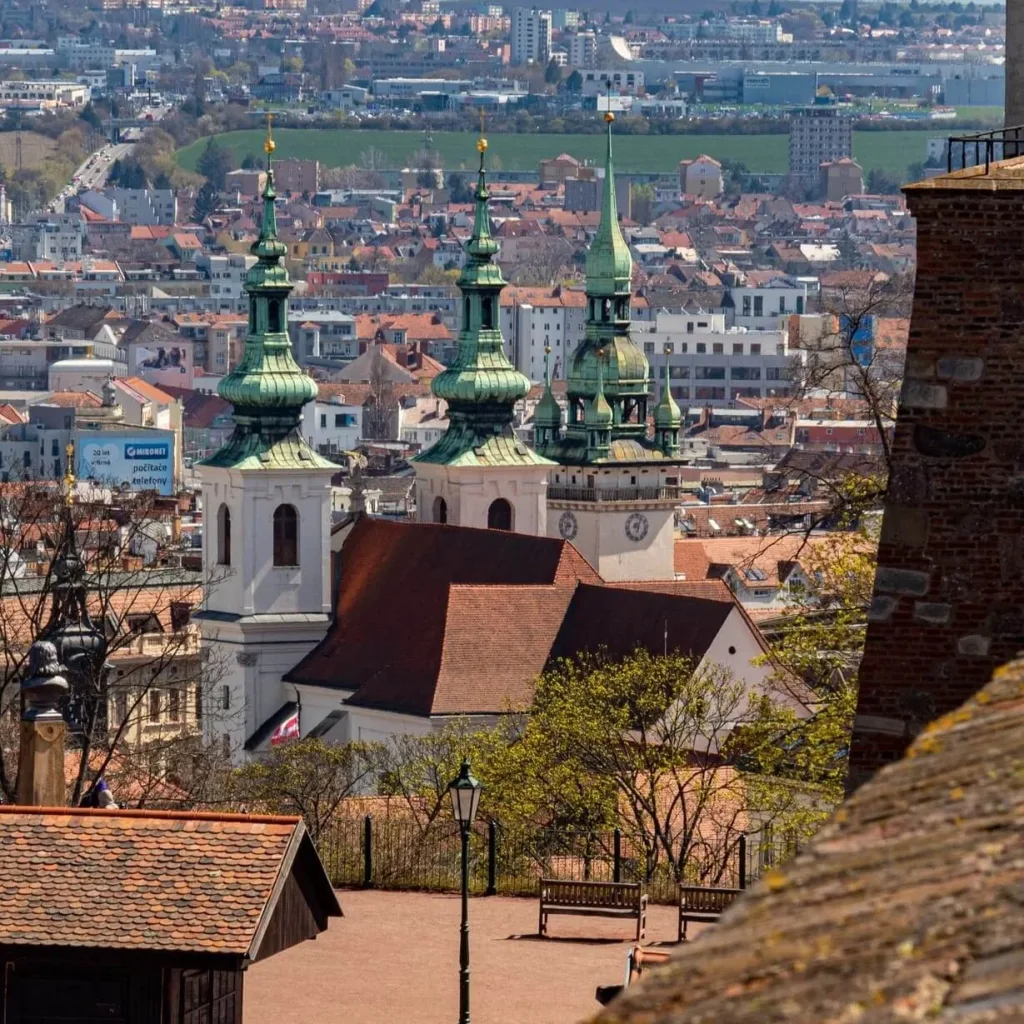 A view over the red rooftops of Brno to the green onion domes of a twin-towered baroque church, seen from a terrace above the old town.