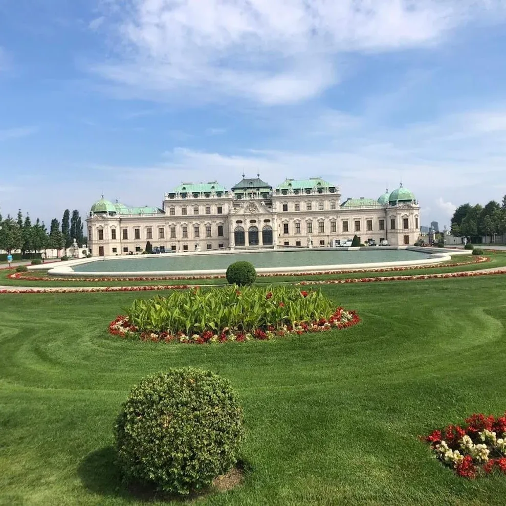 The Upper Belvedere palace in Vienna seen across its formal garden, the long baroque facade and green copper domes mirrored in the reflecting pool.