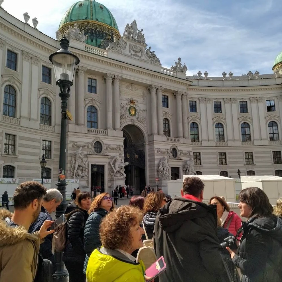 A tour group standing with their guide on Michaelerplatz in Vienna, in front of the green-domed St Michael's Wing of the Hofburg Palace.