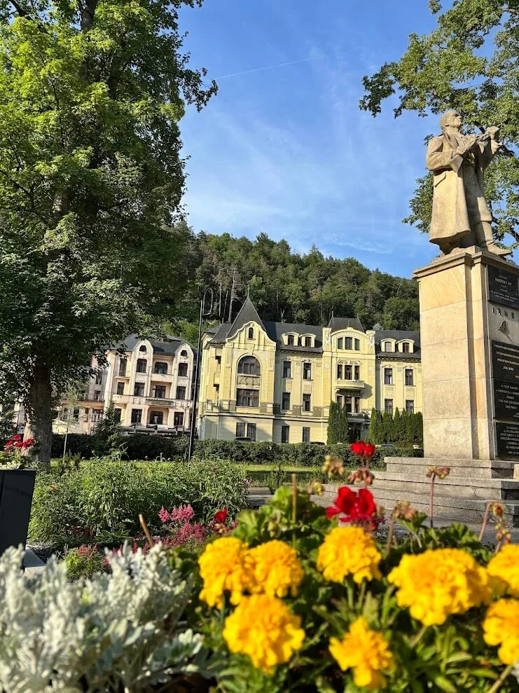 The spa park in Trenčianske Teplice, with a statue on its pedestal, flowerbeds in bloom and a yellow historic spa building behind.