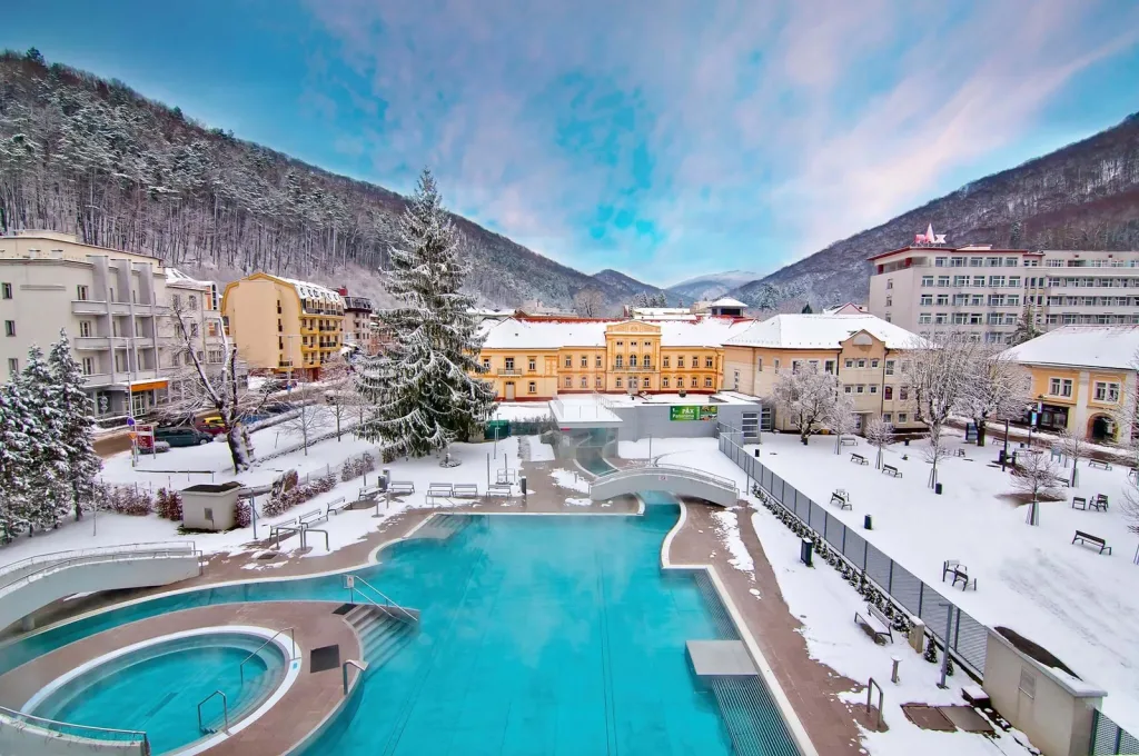 Trenčianske Teplice in winter: the steaming outdoor thermal pool in the spa park, with snow-covered spa buildings, the PAX hotel and wooded hills behind.