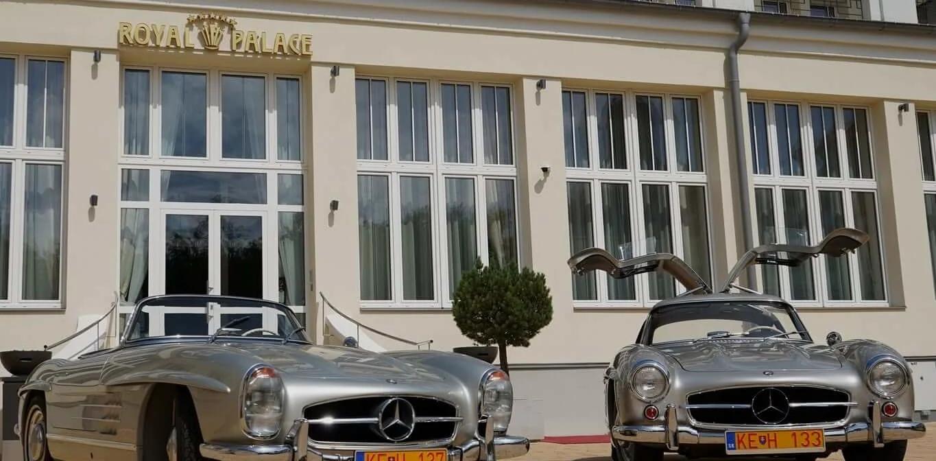 Two classic silver Mercedes-Benz 300 SL sports cars, a roadster and a gullwing coupé, parked outside the Royal Palace hotel in Turčianske Teplice.