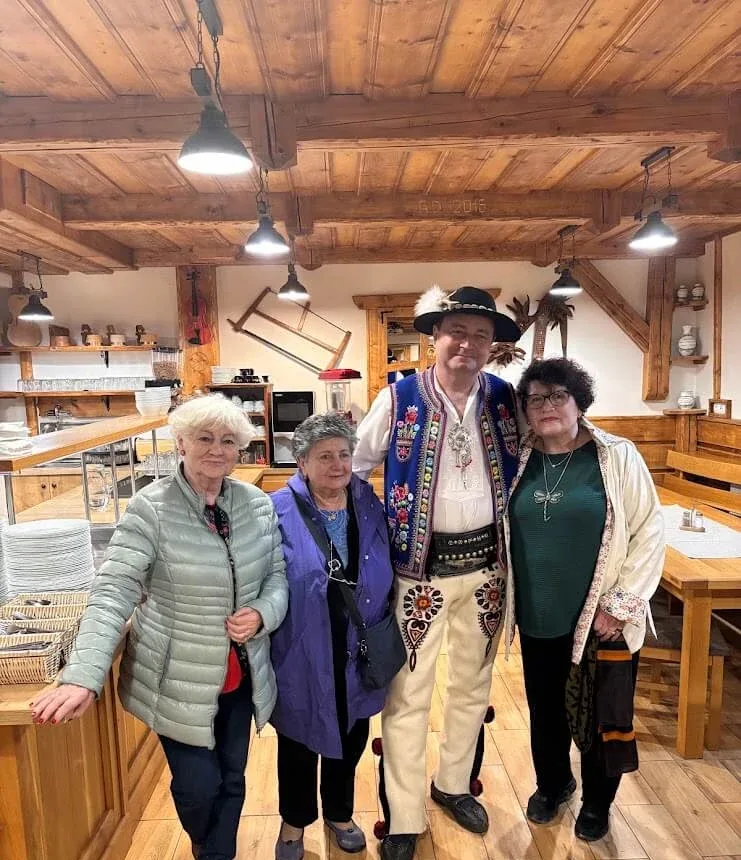 Three visitors with a host in full traditional Slovak dress — feathered hat, embroidered waistcoat and decorated trousers — in a timber-beamed mountain restaurant.