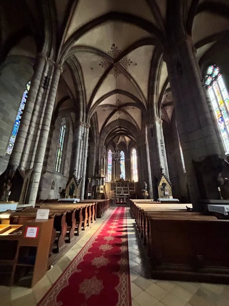 The Gothic nave of the abbey church at Hronsky Benadik, its ribbed vault and stained glass above a red-carpeted aisle.