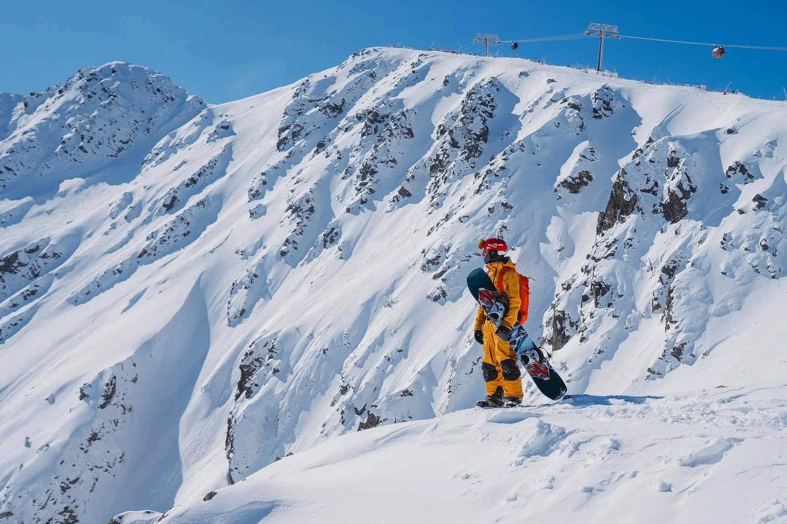Un snowboardeur debout sur une crête enneigée à Jasná, dans les Basses Tatras slovaques, avec une télécabine traversant la crête au-dessus et des sommets enneigés au loin.