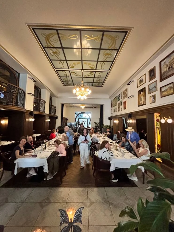 A group at lunch in an Art Nouveau restaurant in Košice, beneath a stained-glass ceiling panel and walls hung with paintings.