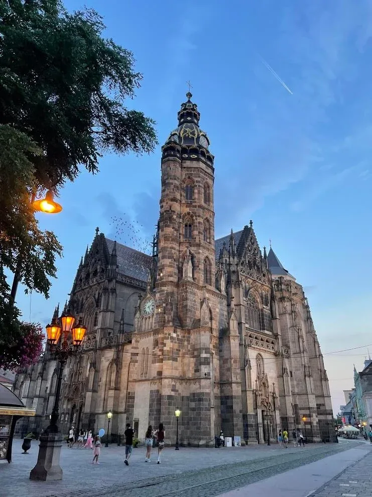 St Elisabeth Cathedral and the Urban Tower in Košice at dusk, the street lamps lit along Hlavná and people walking on the square below.