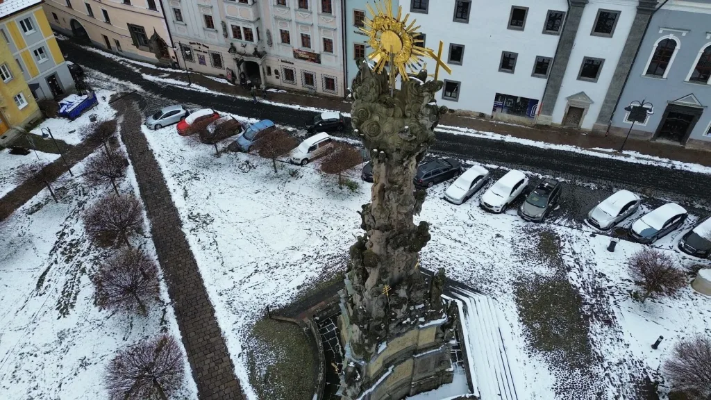 An aerial view of the plague column on Kremnica's main square in winter, snow lying between the parked cars and the painted houses.