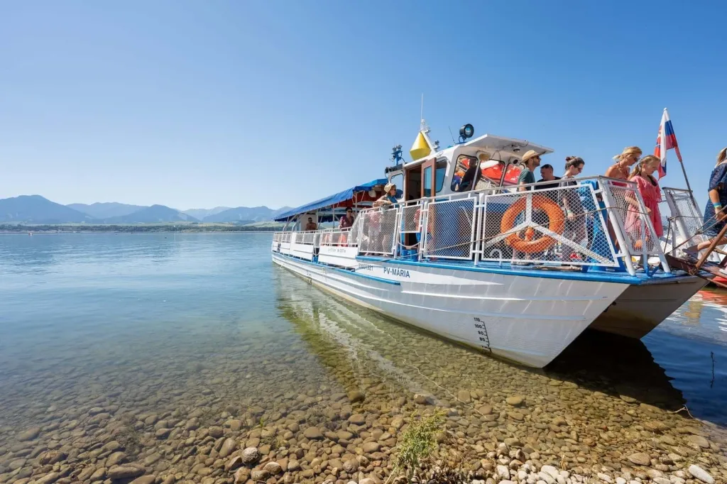 An excursion boat moored at the stony shore of a lake in the Liptov region, passengers on deck and mountains behind.
