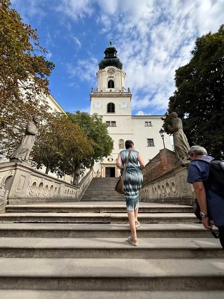 Visitors climbing the balustraded stone stairway up to Nitra Castle, baroque statues flanking the steps and the white clock tower above.