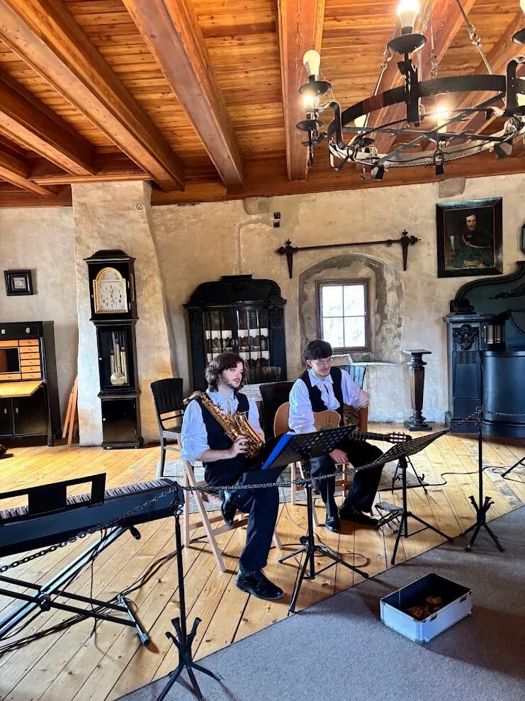 Two musicians with a saxophone and guitar playing in a timber-ceilinged room inside Orava Castle.