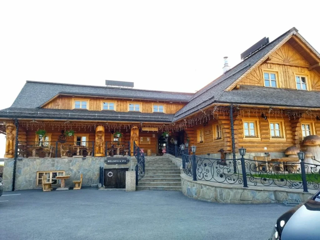 A large traditional log koliba restaurant in Orava, its carved timber facade and stone steps under a bright sky.