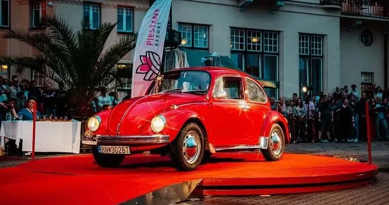 Красный Volkswagen Beetle на красной ковровой дорожке на Винтажном вечере в Пьештянах, за ним наблюдает толпа рядом с баннером Piešťany Rezort.