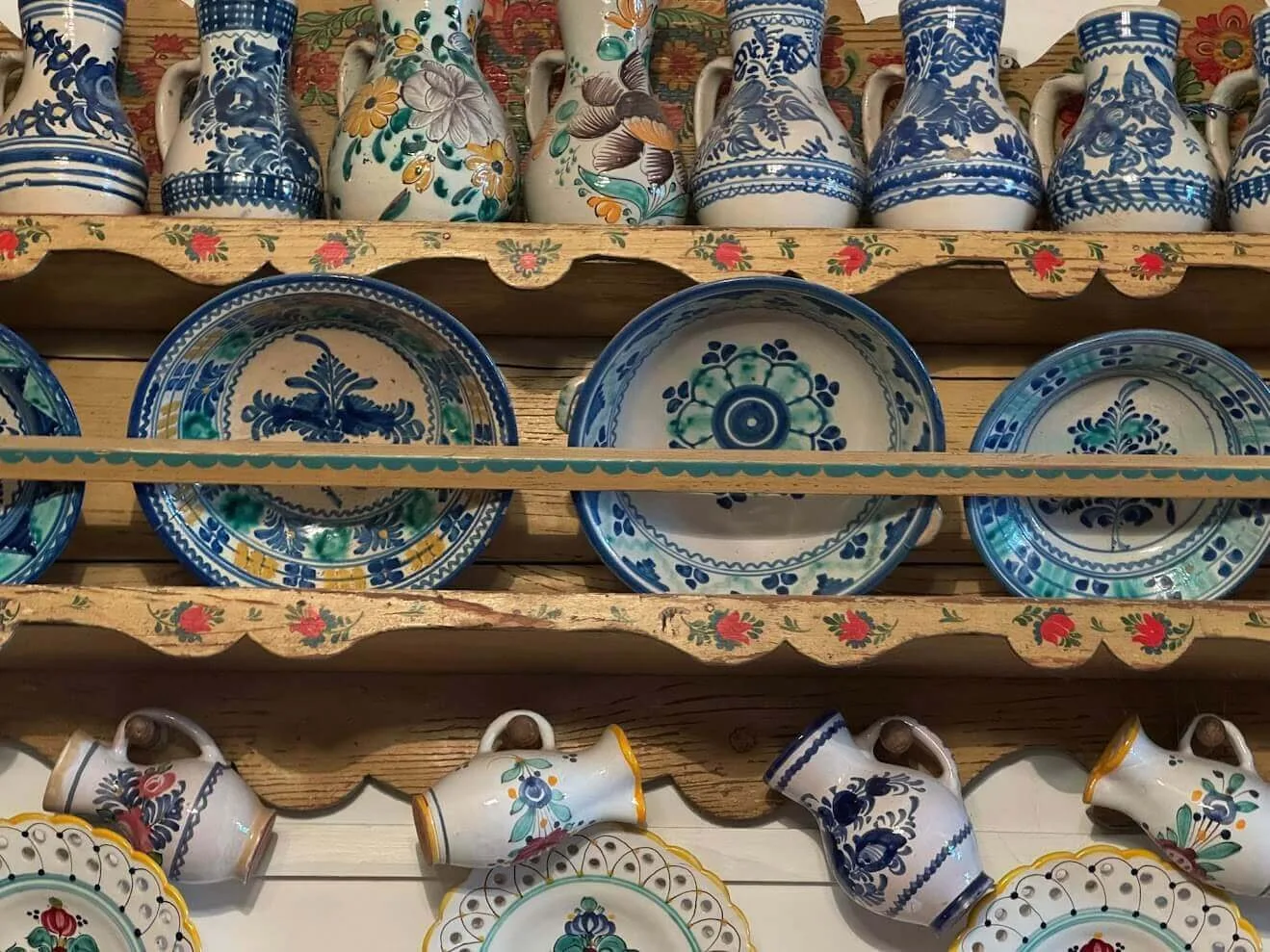 Cerámica eslovaca de mayólica pintada a mano en estantes de madera: jarras, platos y cuencos con motivos azules, verdes y amarillos.