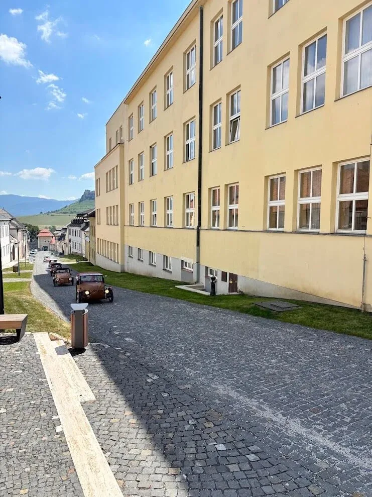 A cobbled street at Spišská Kapitula with vintage cars parked along it and the ruins of Spiš Castle on the horizon.