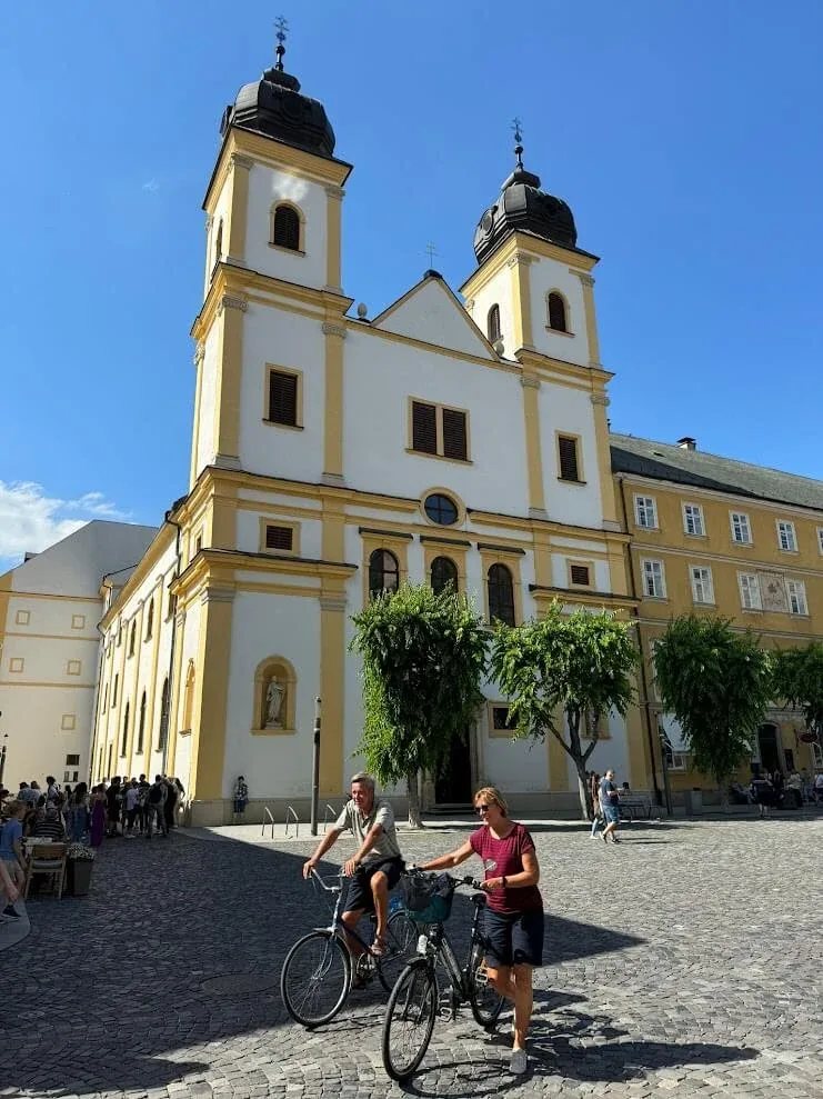 The twin-towered yellow and white Piarist Church on Trenčín's main square, with two cyclists crossing the cobbles in front.