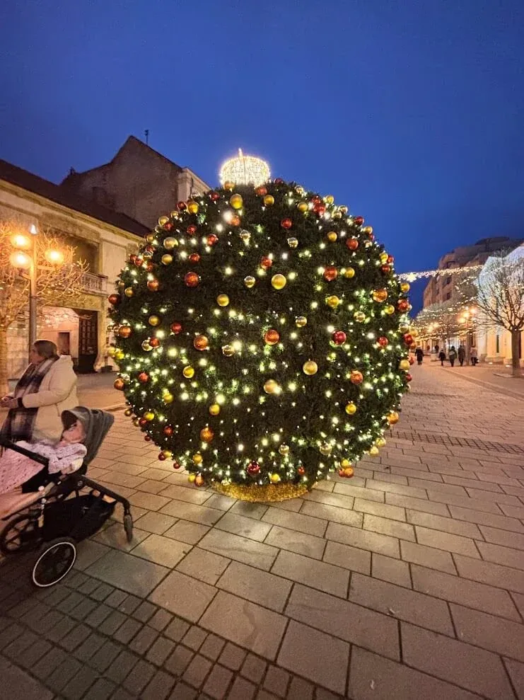 A large spherical Christmas tree covered in lights and baubles standing on Trnava's pedestrian street at dusk.