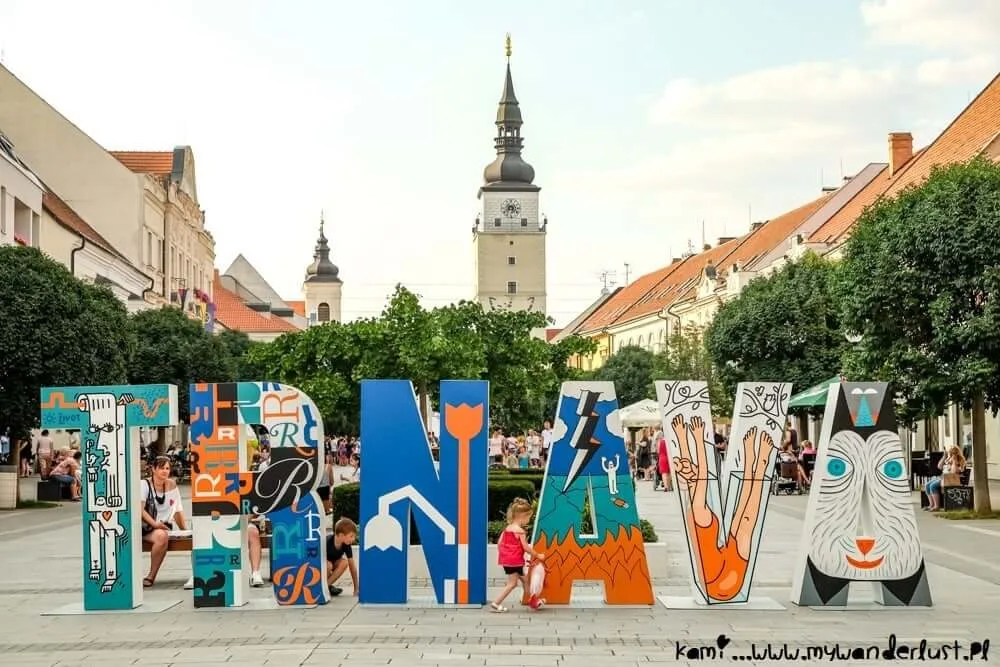 Des enfants jouant parmi les lettres colorées peintes TRNAVA sur la place principale, avec la tour de la ville et les flèches des églises derrière.