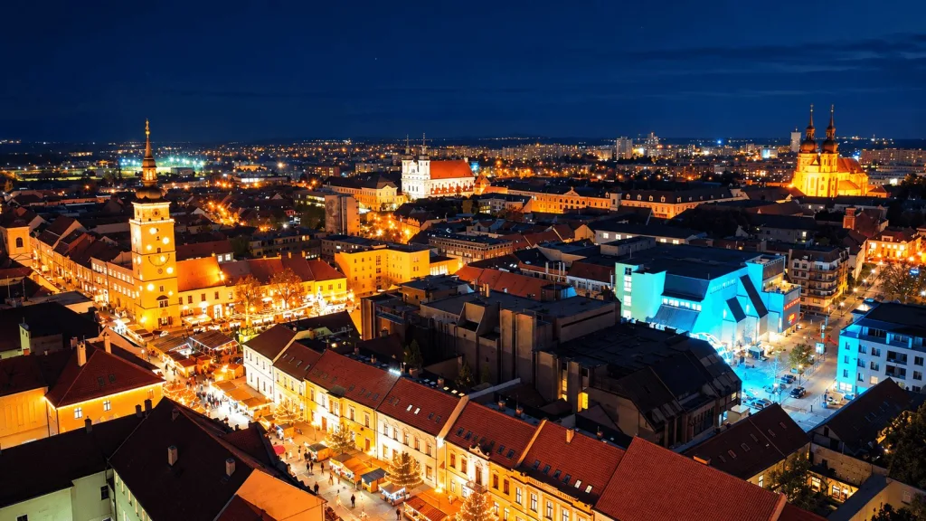 La vieille ville de Trnava vue du ciel de nuit, avec la tour de la ville illuminée, les flèches des églises et les étals du marché sur la place principale.