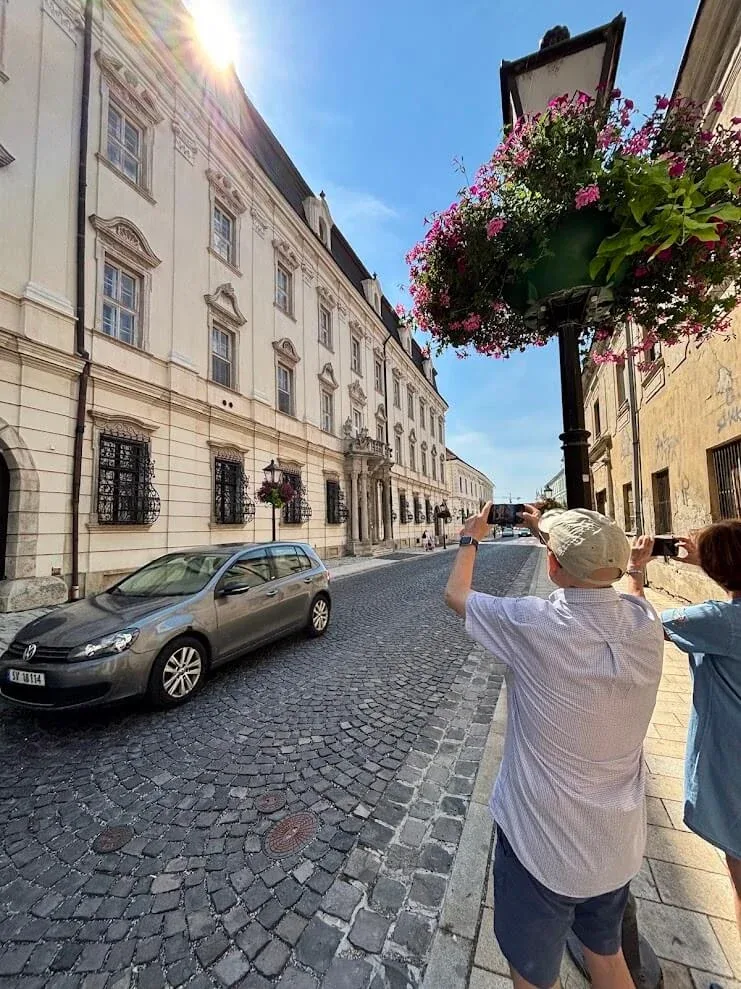 Visitors photographing the buildings along a cobbled street in Trnava, with hanging baskets of pink flowers overhead.