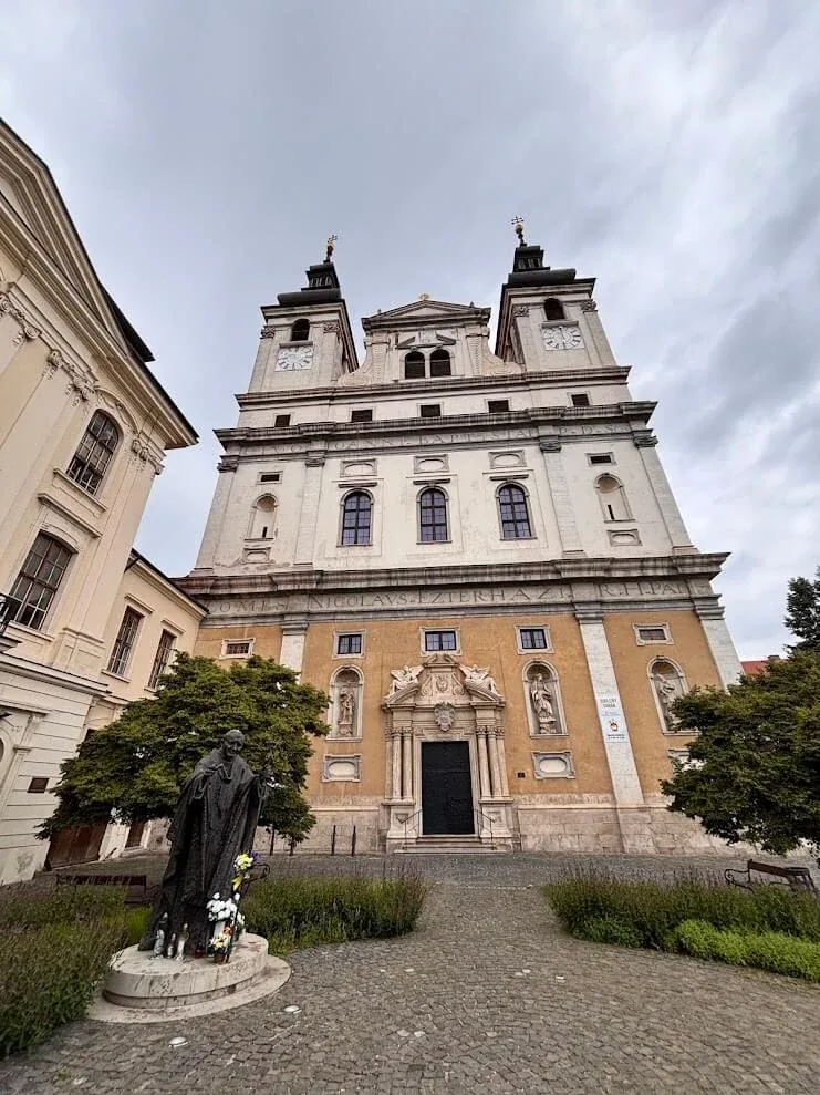 The twin-towered baroque facade of St John the Baptist Cathedral in Trnava, with a bronze statue standing on the square in front of it.