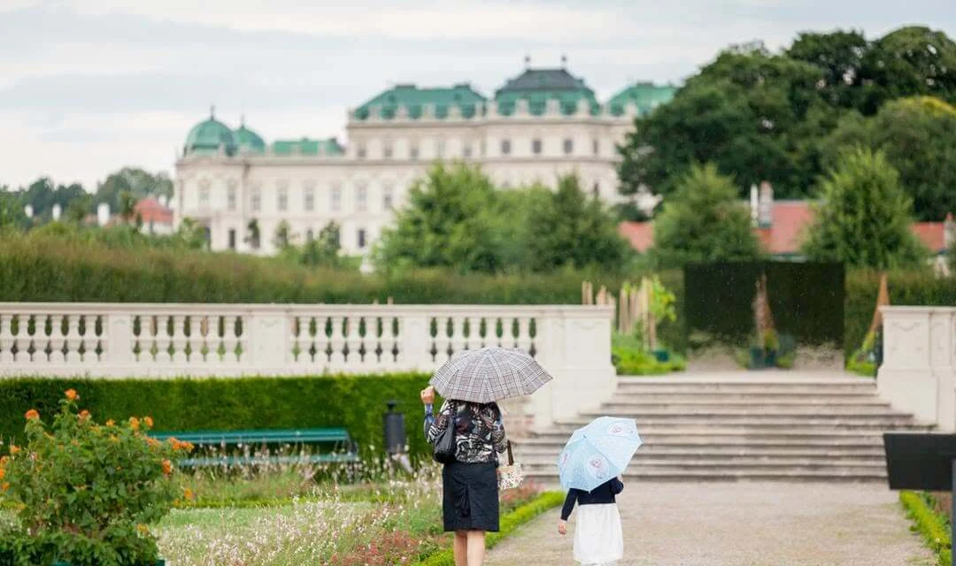 A woman and a child with umbrellas walking through the Belvedere gardens in Vienna, with the Upper Belvedere palace and its green domes beyond the balustrade.