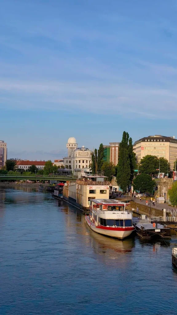 Riverboats moored on the Danube Canal in Vienna at evening, with the domed Urania observatory on the embankment behind.