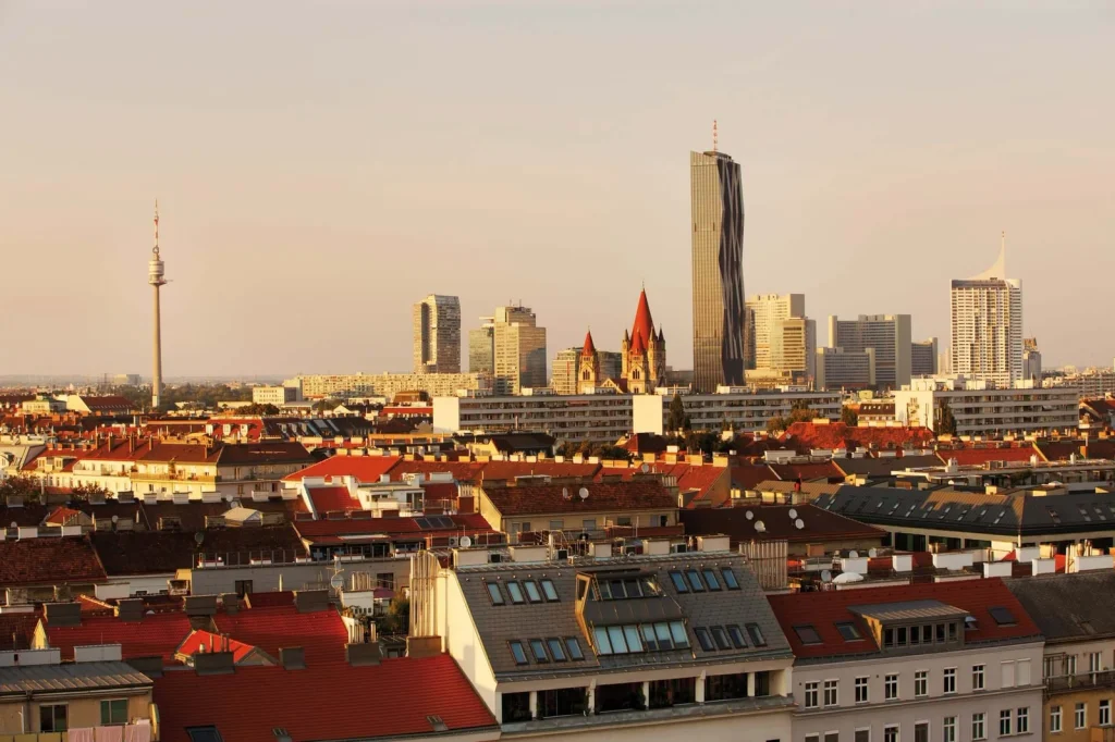 The Vienna skyline at sunset over red rooftops, with the Danube Tower, the DC Tower and a red-spired church rising beyond.