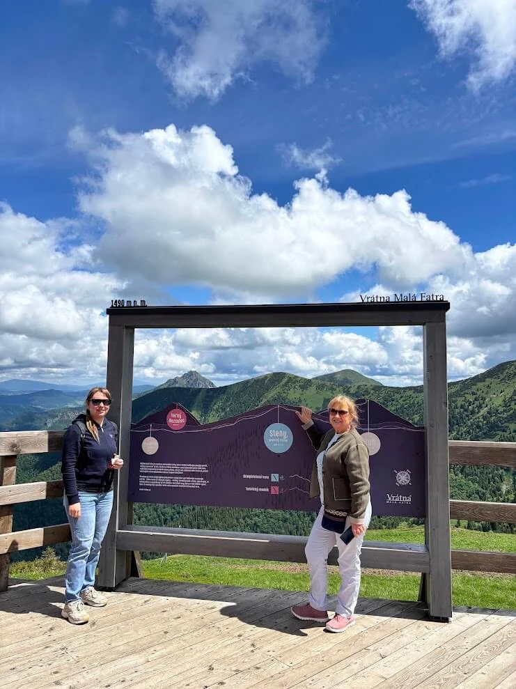 Two visitors at the Vrátna Malá Fatra viewpoint panel at 1,490 m, with the ridge and the peak of Veľký Rozsutec behind.