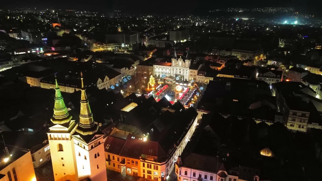 An aerial night view of Žilina, with the Christmas market lit up on Mariánske Square and the floodlit church towers in the foreground.