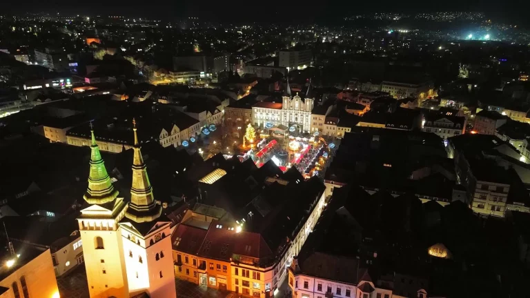 An aerial night view of Žilina, with the Christmas market lit up on Mariánske Square and the floodlit church towers in the foreground.