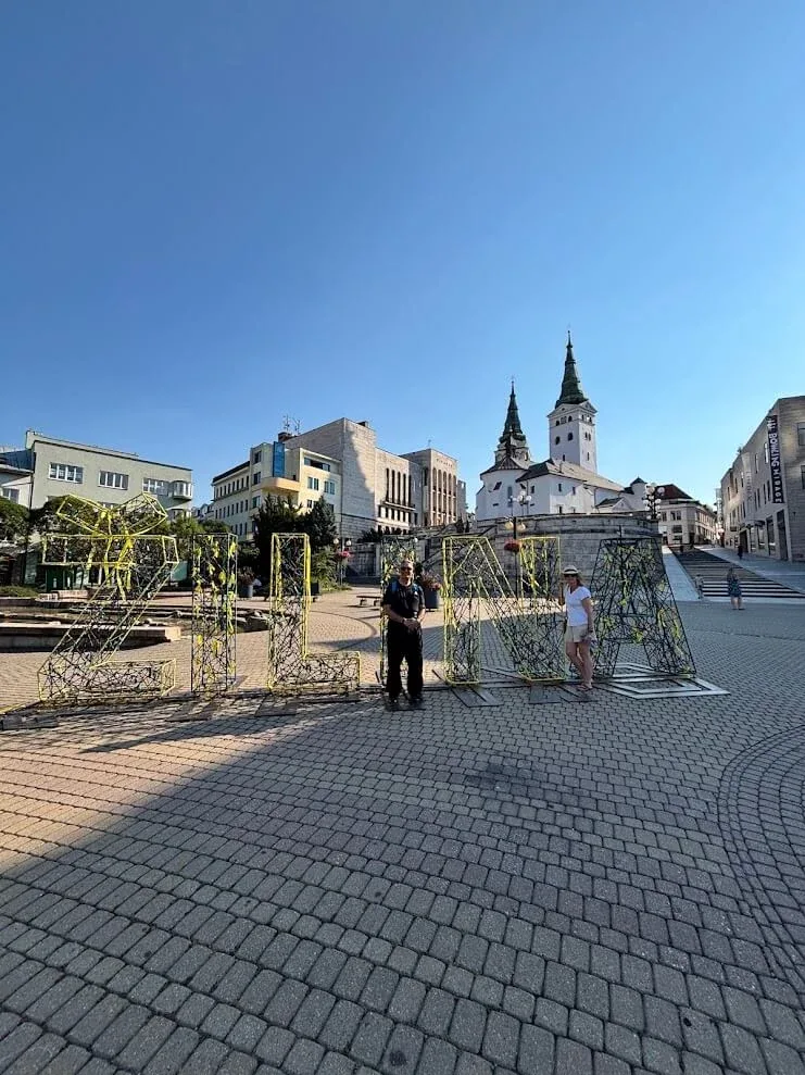 Des visiteurs à côté des lettres ZILINA sur la place principale, avec les deux tours de l'église paroissiale en arrière-plan.