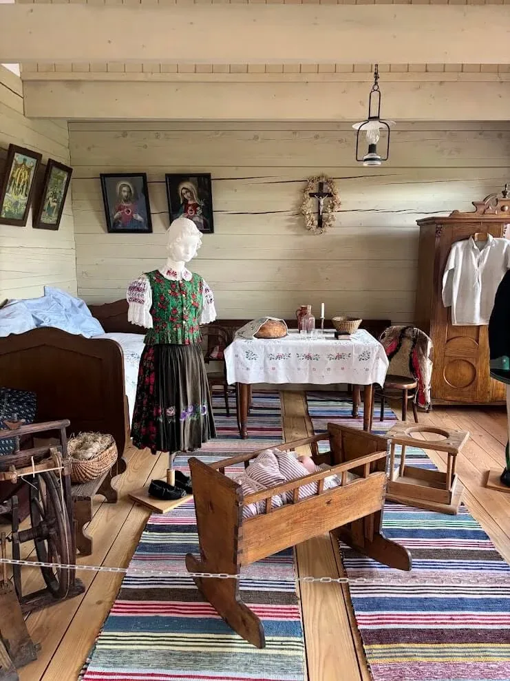 A furnished cottage room at the open-air Museum of Orava Village in Zuberec, with a folk costume, a wooden cradle and a spinning wheel.