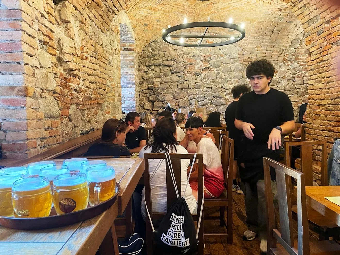 A brick-vaulted cellar beer house in Bratislava, a tray of half-litre lager tankards in the foreground and guests at long wooden tables under the arches beyond.