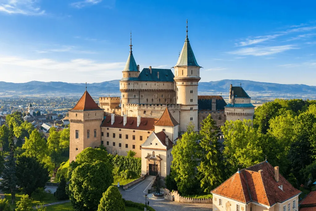 Bojnice Castle from the air, its turrets and green roofs rising out of the park, with the hills of the Upper Nitra valley beyond.