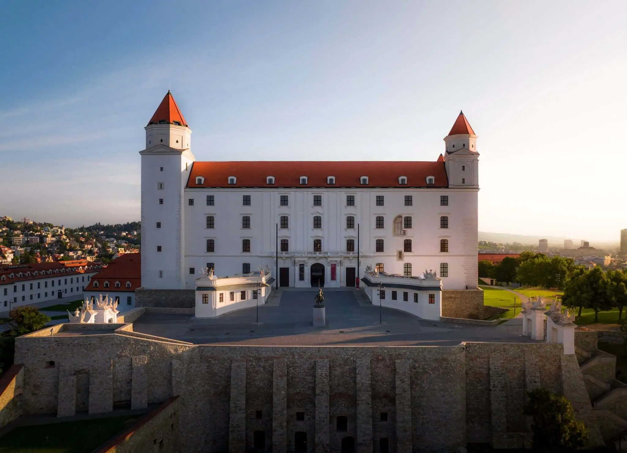 Bratislava Castle in the early morning light, its white walls, red roof and four corner towers standing above the stone ramparts, with the city and the hills beyond.