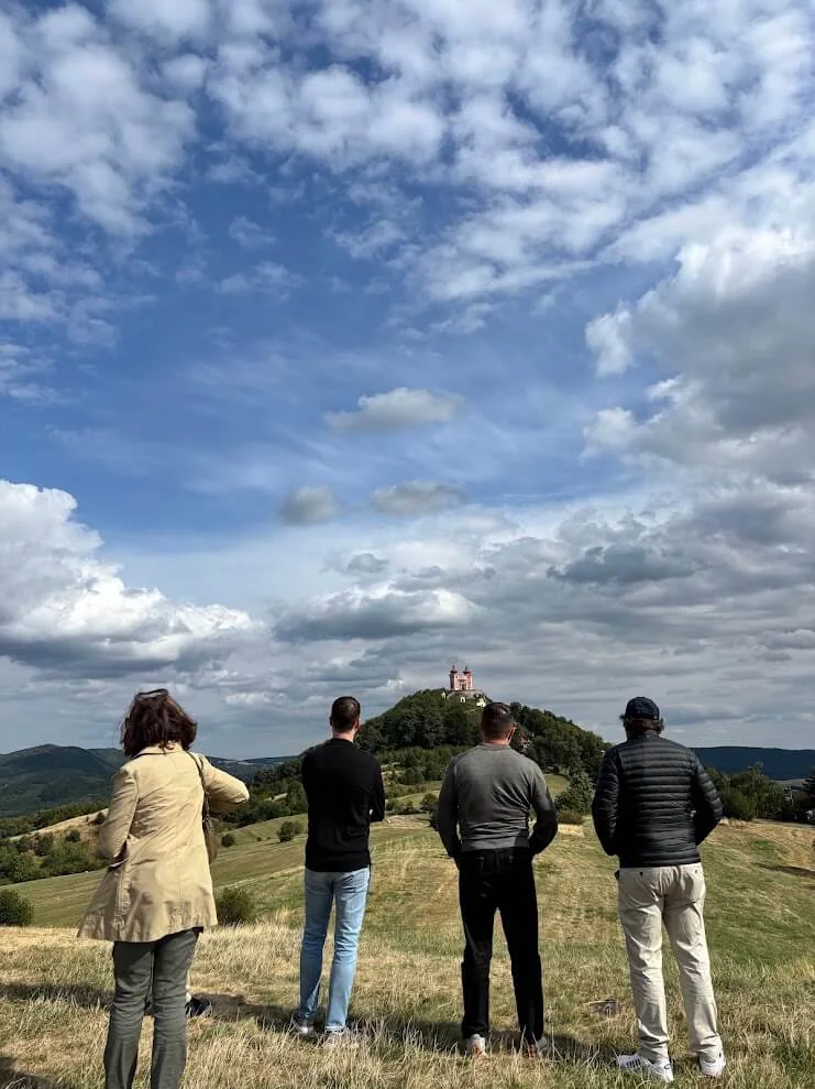 Four visitors looking across the fields to a baroque calvary standing on a conical hill under a wide sky.