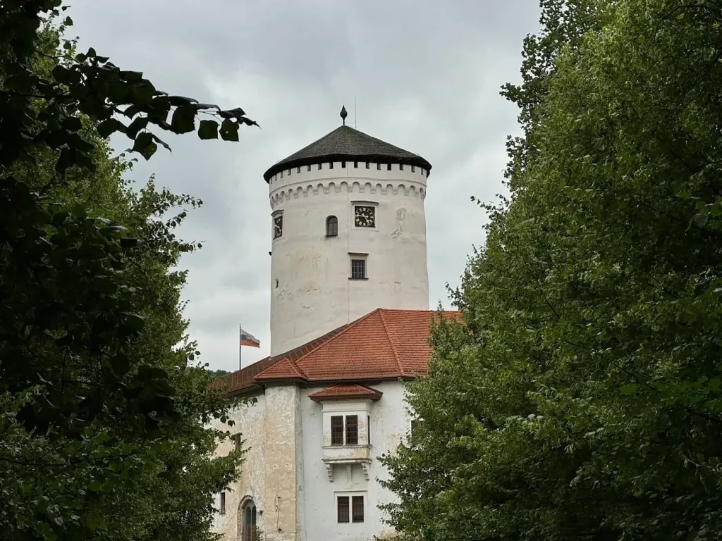 The white round clock tower of Budatín Castle rising above its red-tiled roof, framed by summer trees.