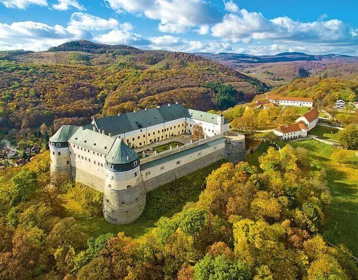 Červený Kameň Castle from the air in autumn, its white walls and round bastions standing above the wooded hills of the Small Carpathians.