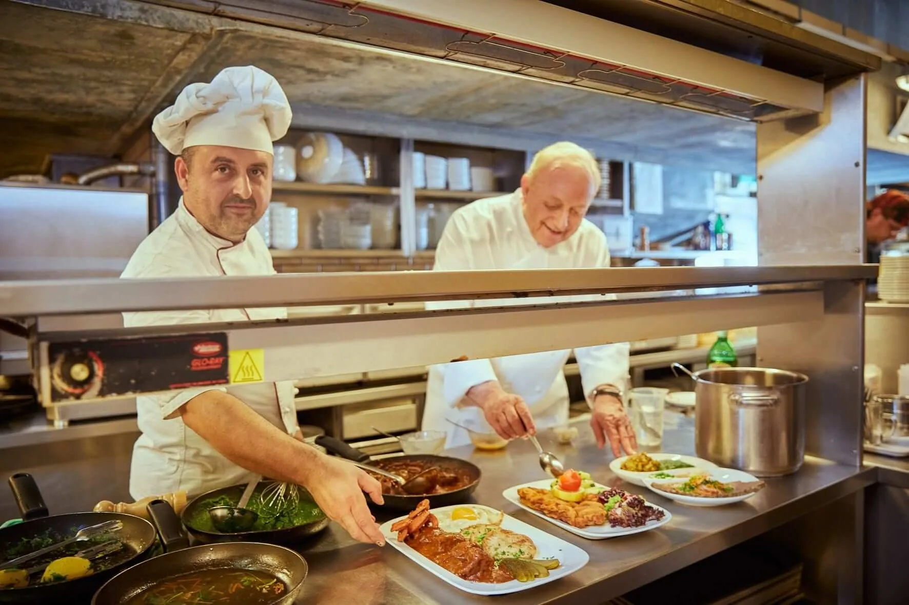 Two chefs at the pass in the kitchen of 12A Keller in Vienna, plating Austrian dishes — schnitzel, goulash with a fried egg, and boiled beef with chive sauce — for a group lunch.