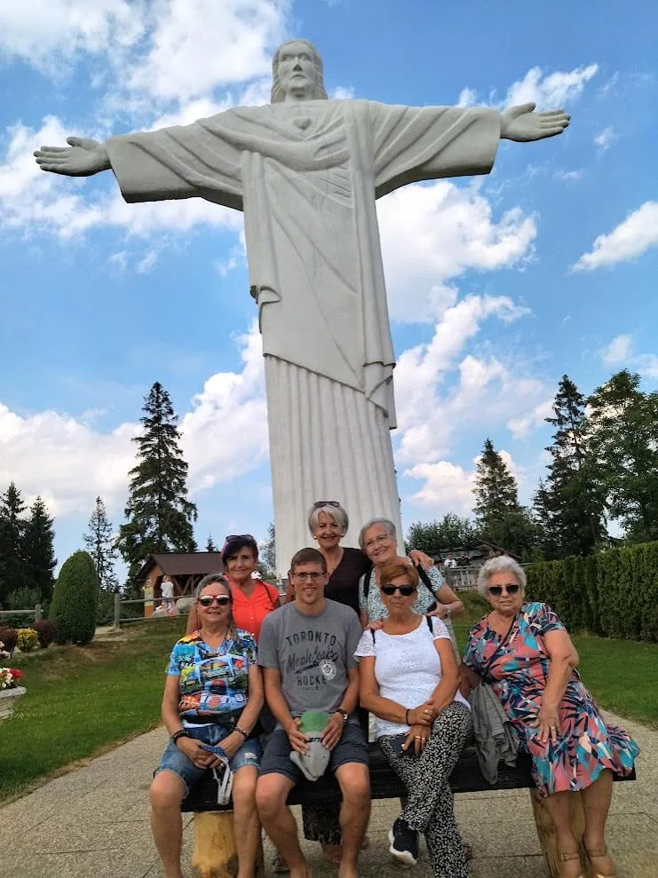 A group of visitors sitting and standing on a bench beneath a tall white statue of Christ with outstretched arms, spruce trees and summer sky behind.