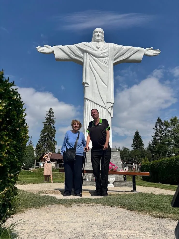 Two visitors standing in front of a tall white statue of Christ with outstretched arms, in a garden under a blue sky.