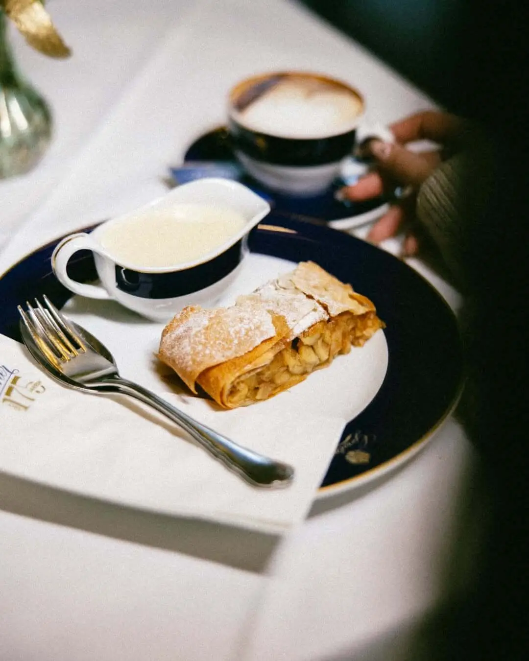 A slice of apple strudel dusted with icing sugar on a dark blue and gold plate at Gerstner in Vienna, served with a jug of vanilla sauce, a melange coffee on the white linen beside it.