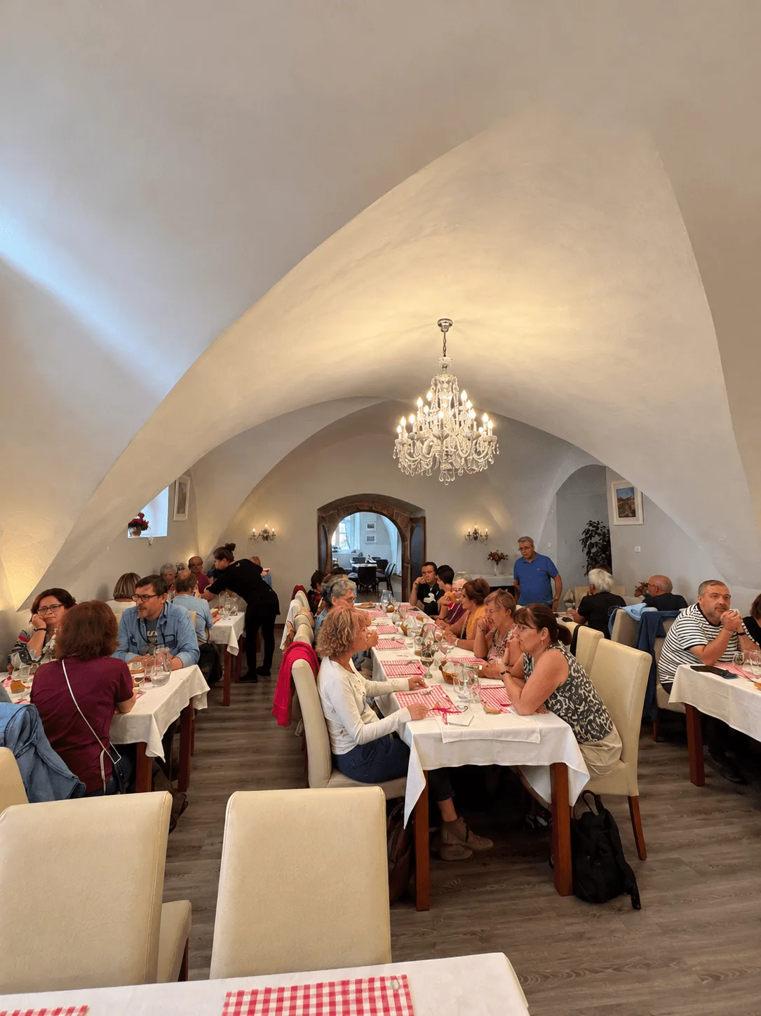 Guests at lunch under the tall white vaulted ceiling of the Galéria restaurant in Banská Štiavnica, red-and-white checked napkins on the white linen and a crystal chandelier hanging from the vault.