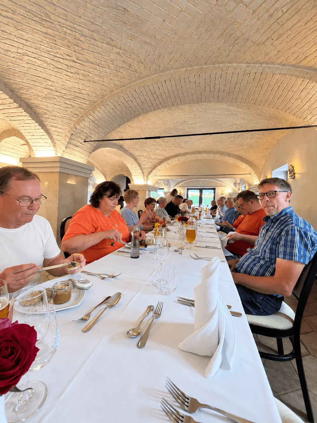 A tour group at a long table laid with white linen, silver and folded napkins under a white-painted vaulted brick ceiling, part-way through a two-course group lunch.