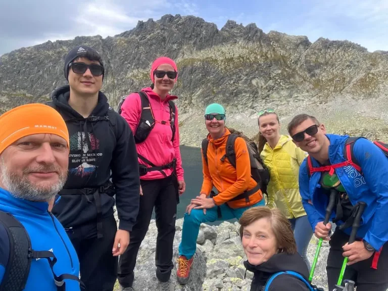A walking group gathered on the rocks beside a dark mountain tarn in the High Tatras, a jagged ridge above them.