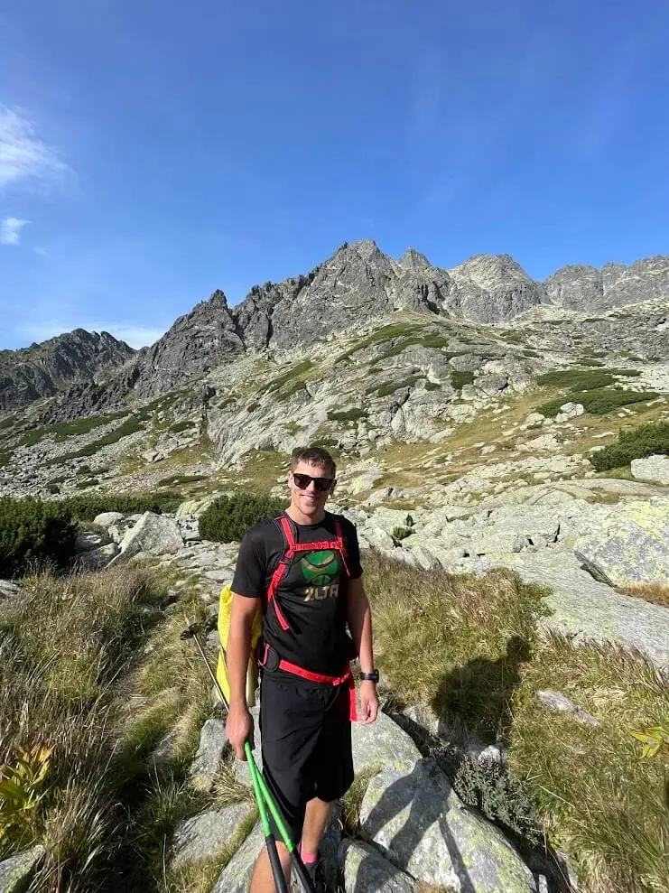 A walker with trekking poles on a boulder-strewn trail in the High Tatras, jagged granite peaks rising behind.